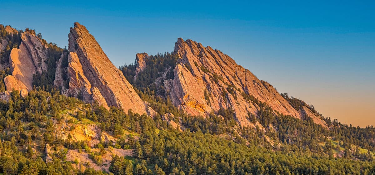 The Flatirons rock formations near Boulder, Colorado, glowing in warm light above an evergreen forest in the Mountain West region.