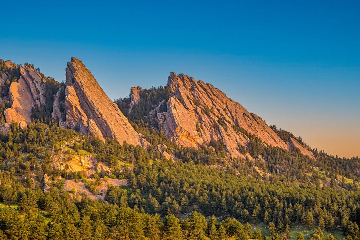 The Flatirons rock formations near Boulder, Colorado, glowing in warm light above an evergreen forest in the Mountain West region.